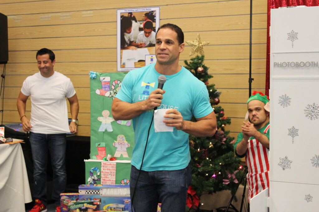 A man in a teal T-shirt speaking into a microphone beside a Christmas tree at a toy drive