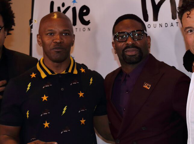 DJ Irie and another man in a star-print shirt posing in front of an Irie Foundation backdrop