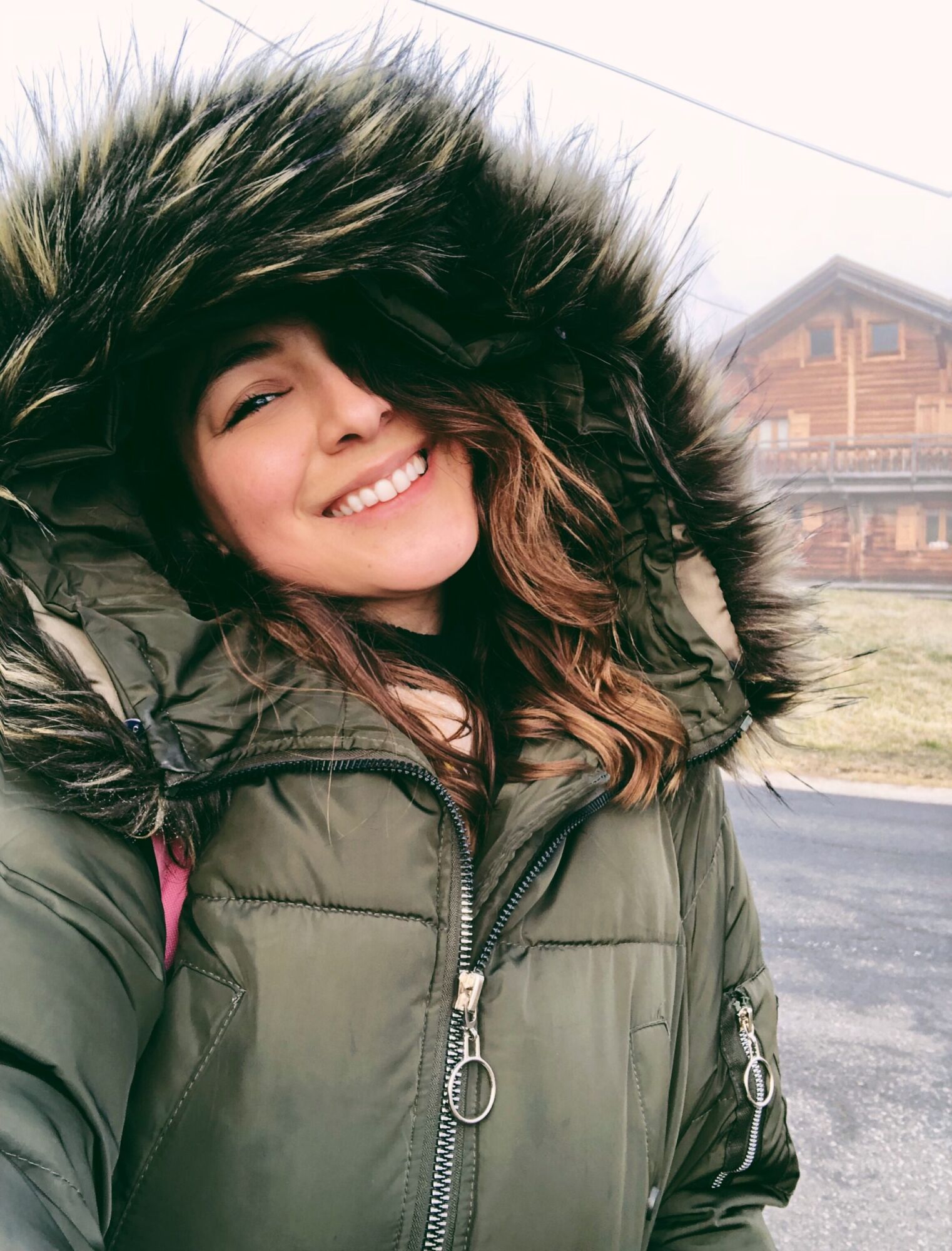 Cata laughing in a khaki parka with a fur-trimmed hood, a wooden chalet behind her
