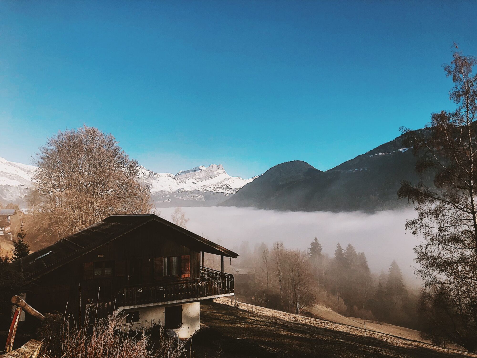 A dark wooden chalet on a hillside above a valley filled with low cloud, bare trees and snowy peaks beyond