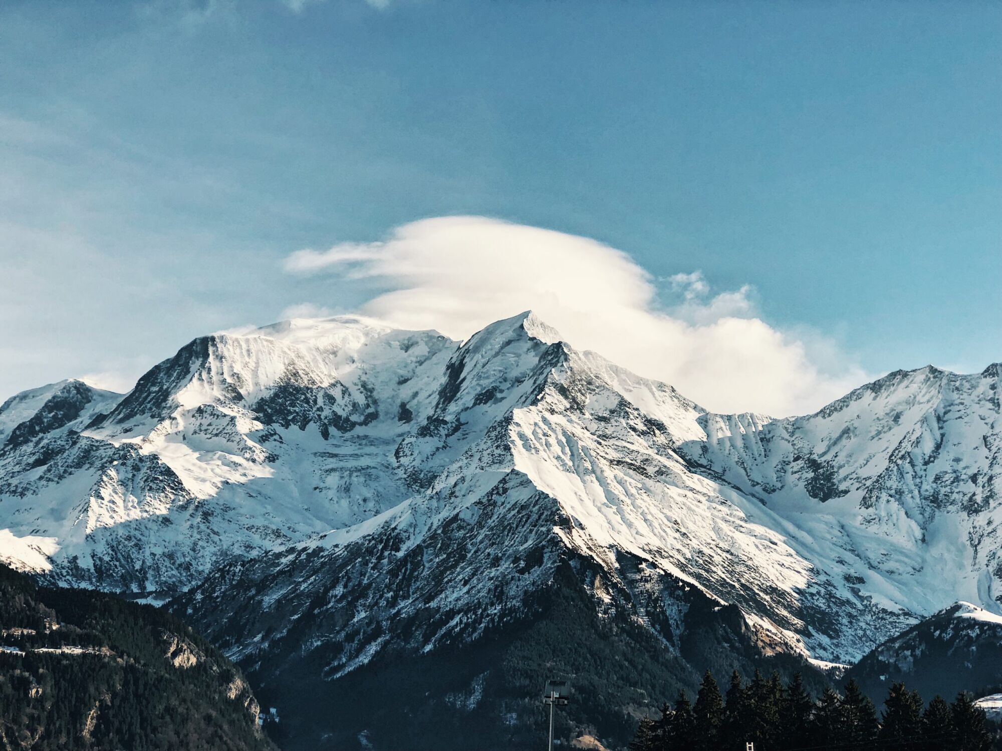 The snow-covered mountains surrounding Saint Gervais-les-Bains under a lenticular cloud, forested slopes below and clear blue sky above
