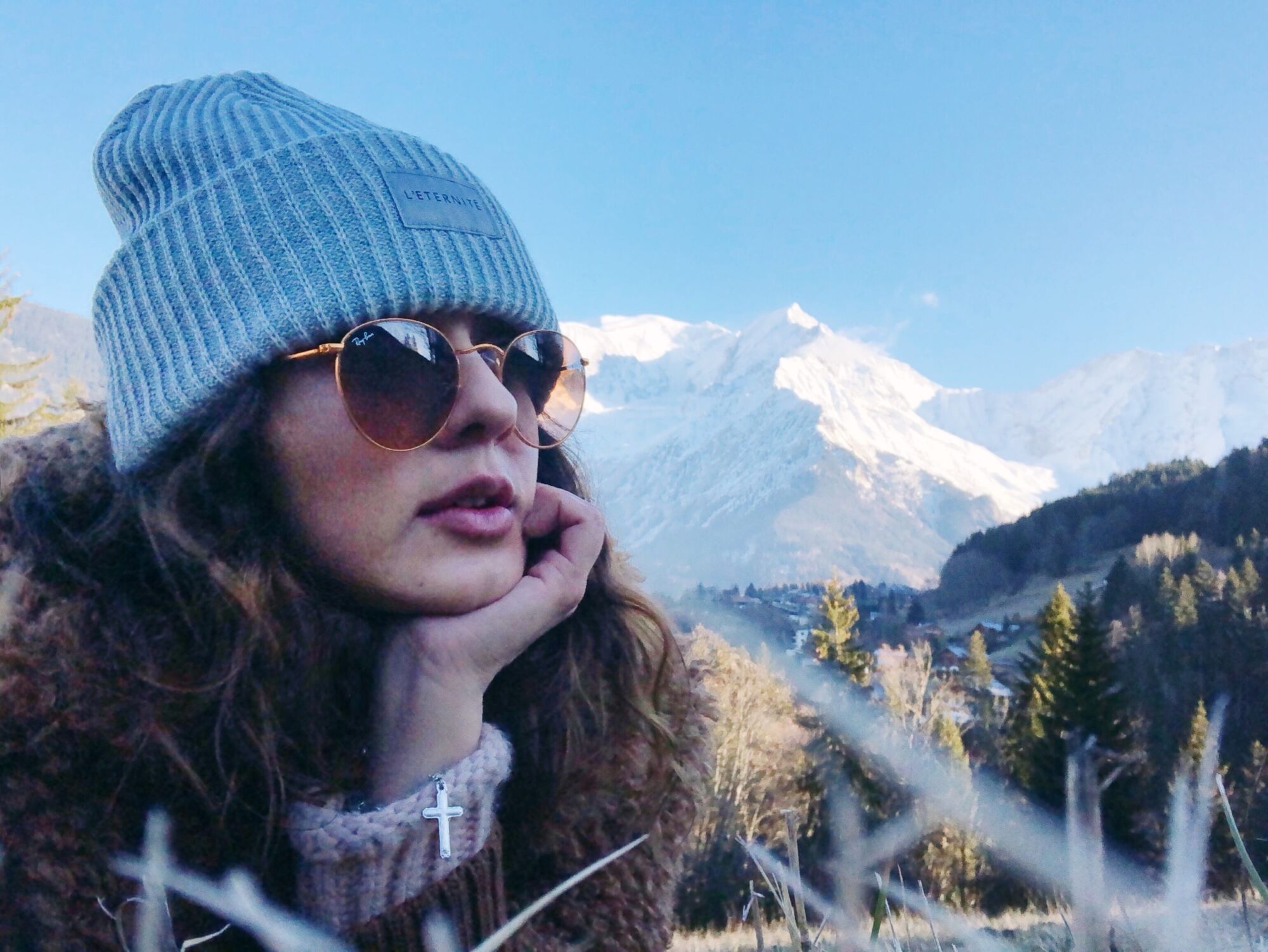 Cata in a grey beanie and sunglasses resting her chin on her hand, the snow-covered mountains surrounding Saint Gervais-les-Bains behind her