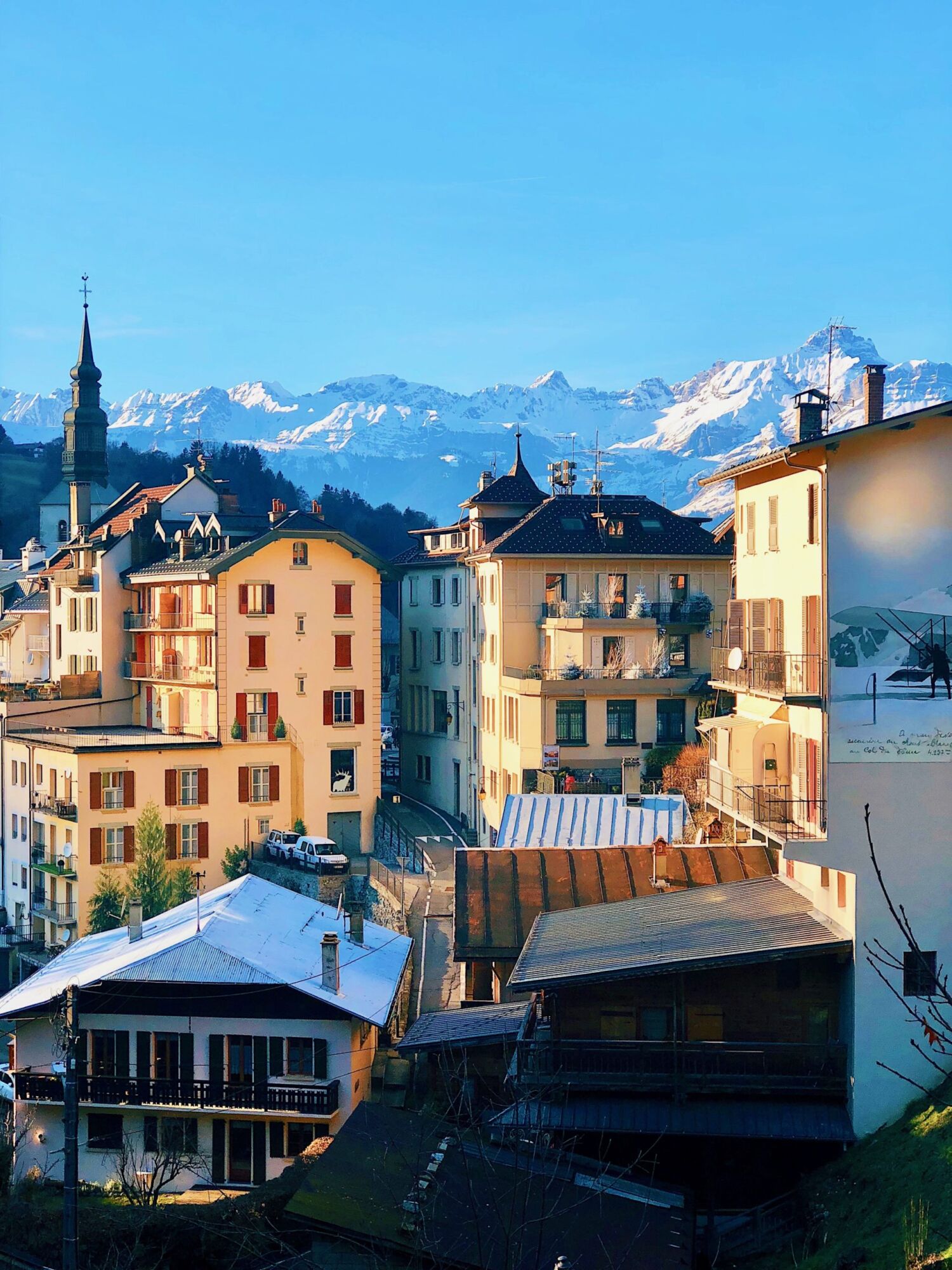 Cream and pastel buildings with a church spire in Saint Gervais-les-Bains, snow on the rooftops and snow-covered peaks rising behind the town