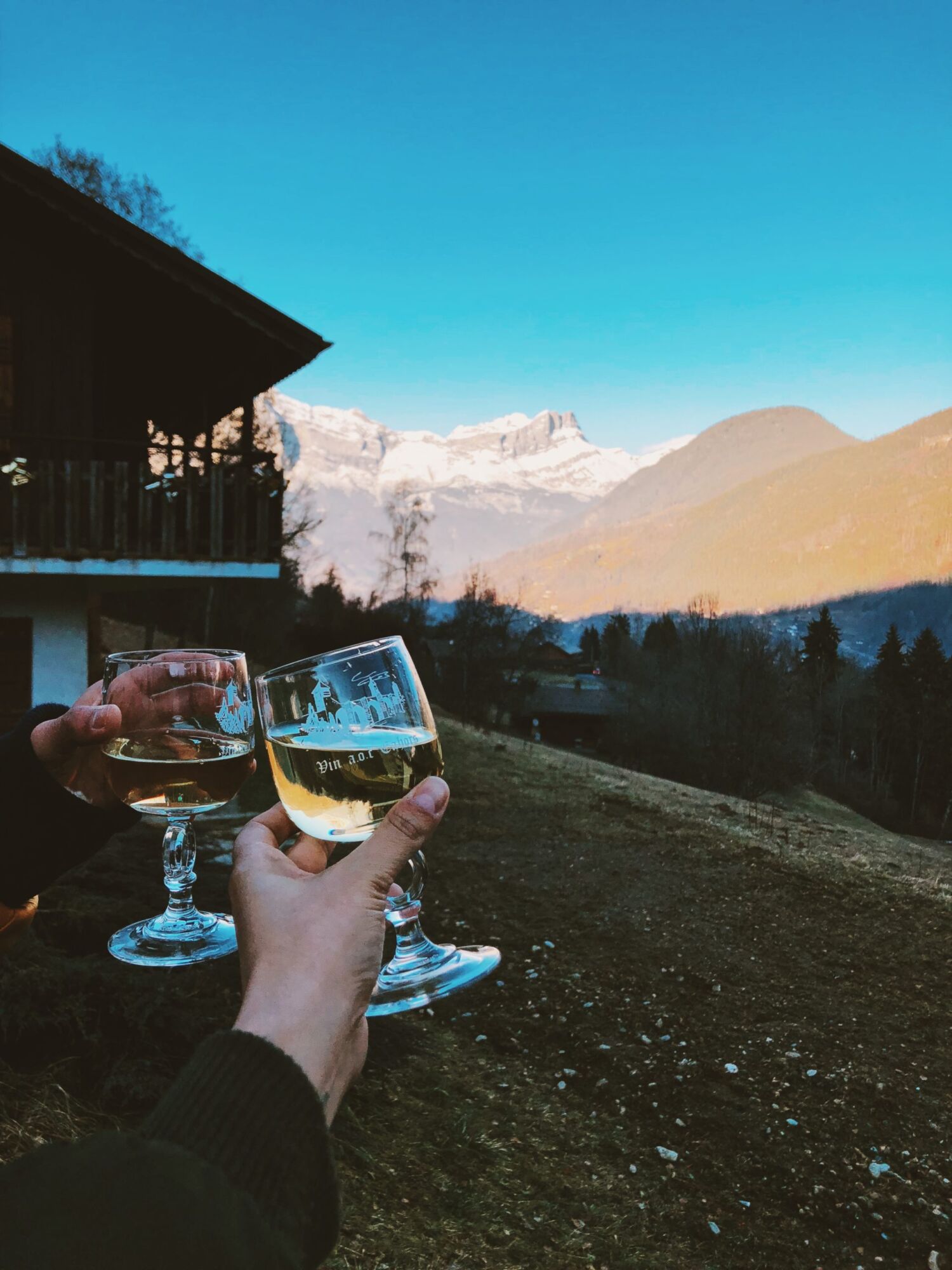 Two glasses of white wine held up against a chalet balcony and snow-covered mountains at dusk
