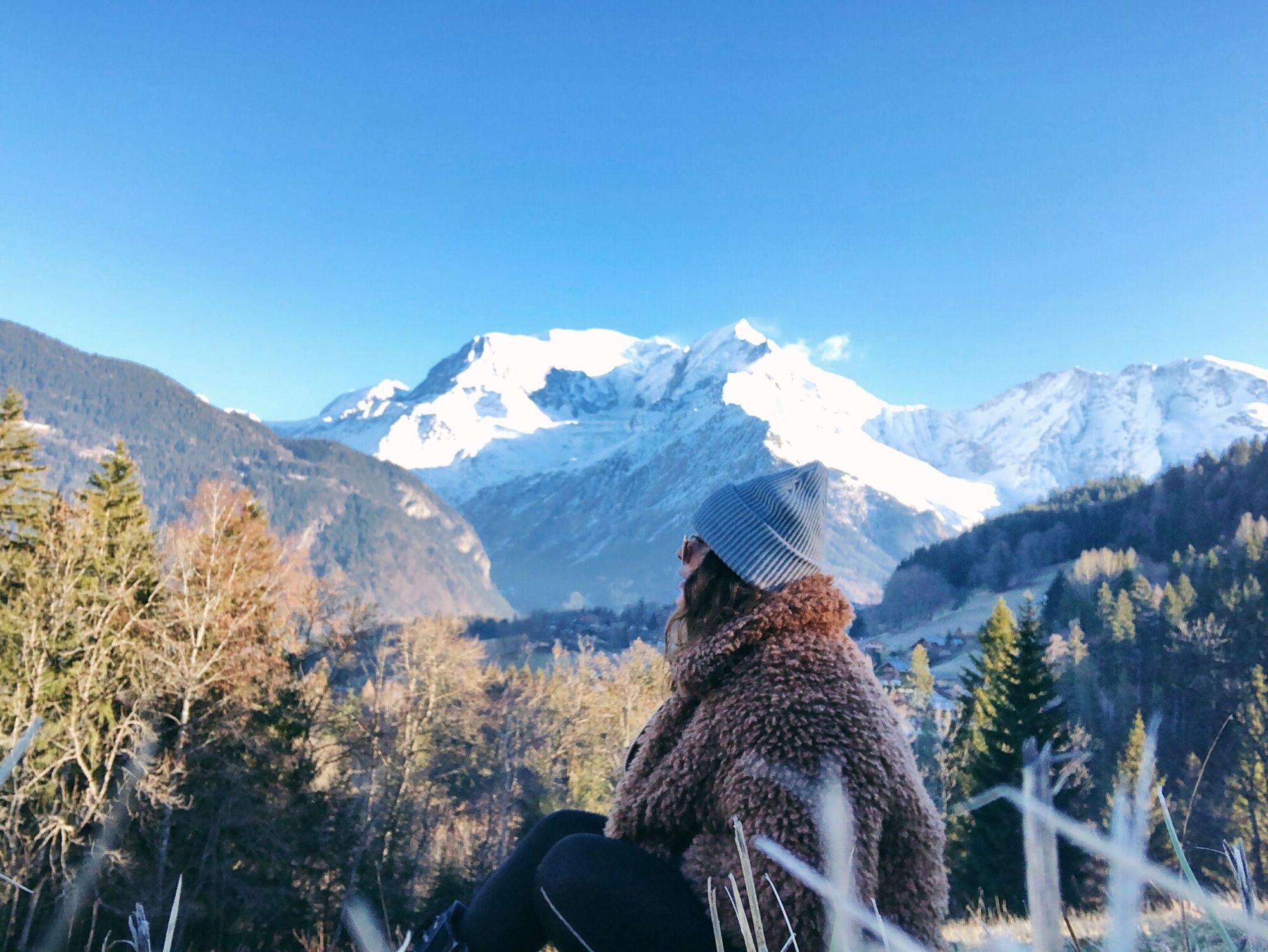 Cata in a faux-fur coat looking out towards the snow-covered mountains surrounding Saint Gervais-les-Bains, pine trees below