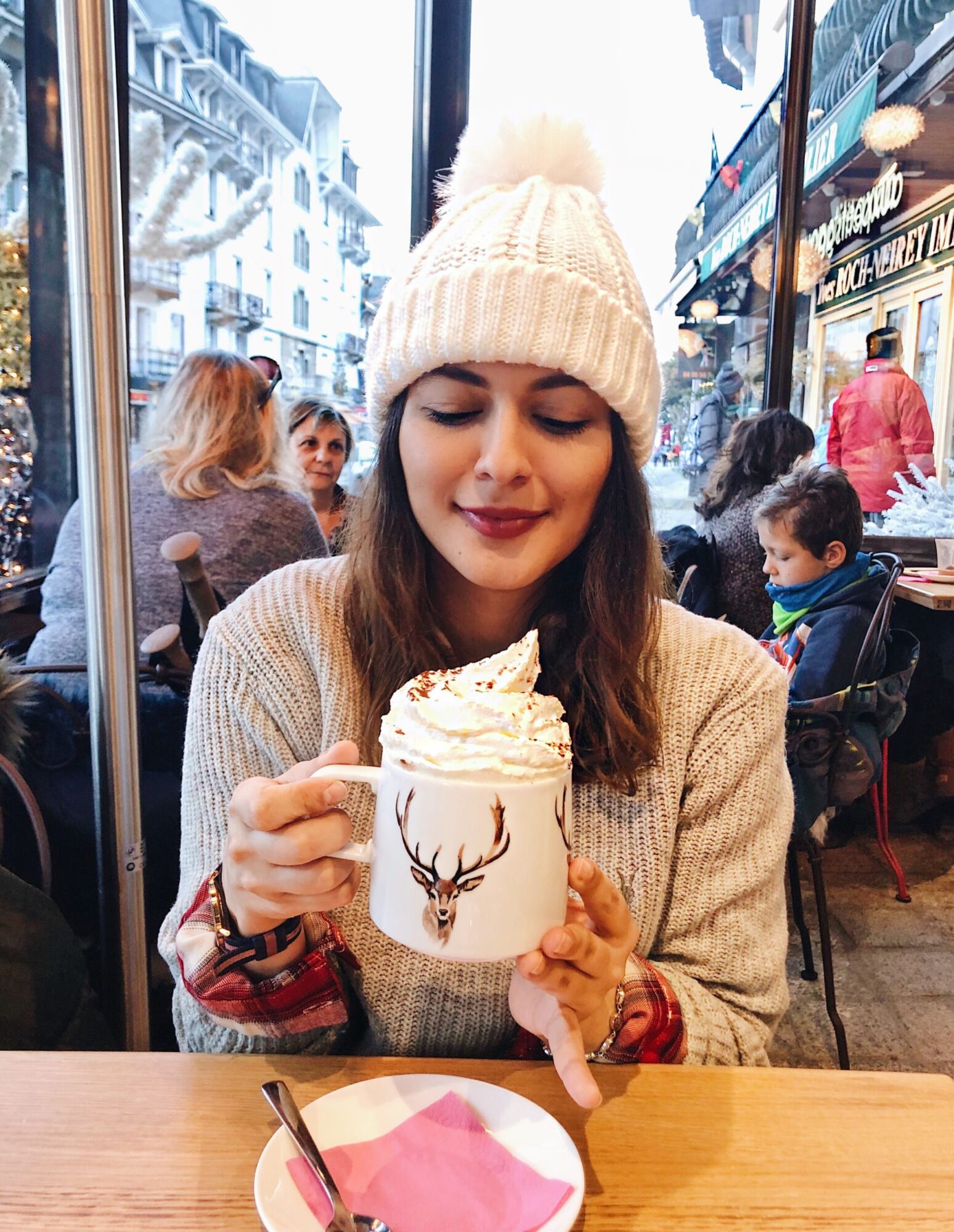 Cata in a white bobble hat holding a tall glass mug of hot chocolate topped with cream, at a café window