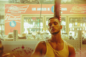 C. Tangana in a white vest under warm light in front of a bar lined with Budweiser signs