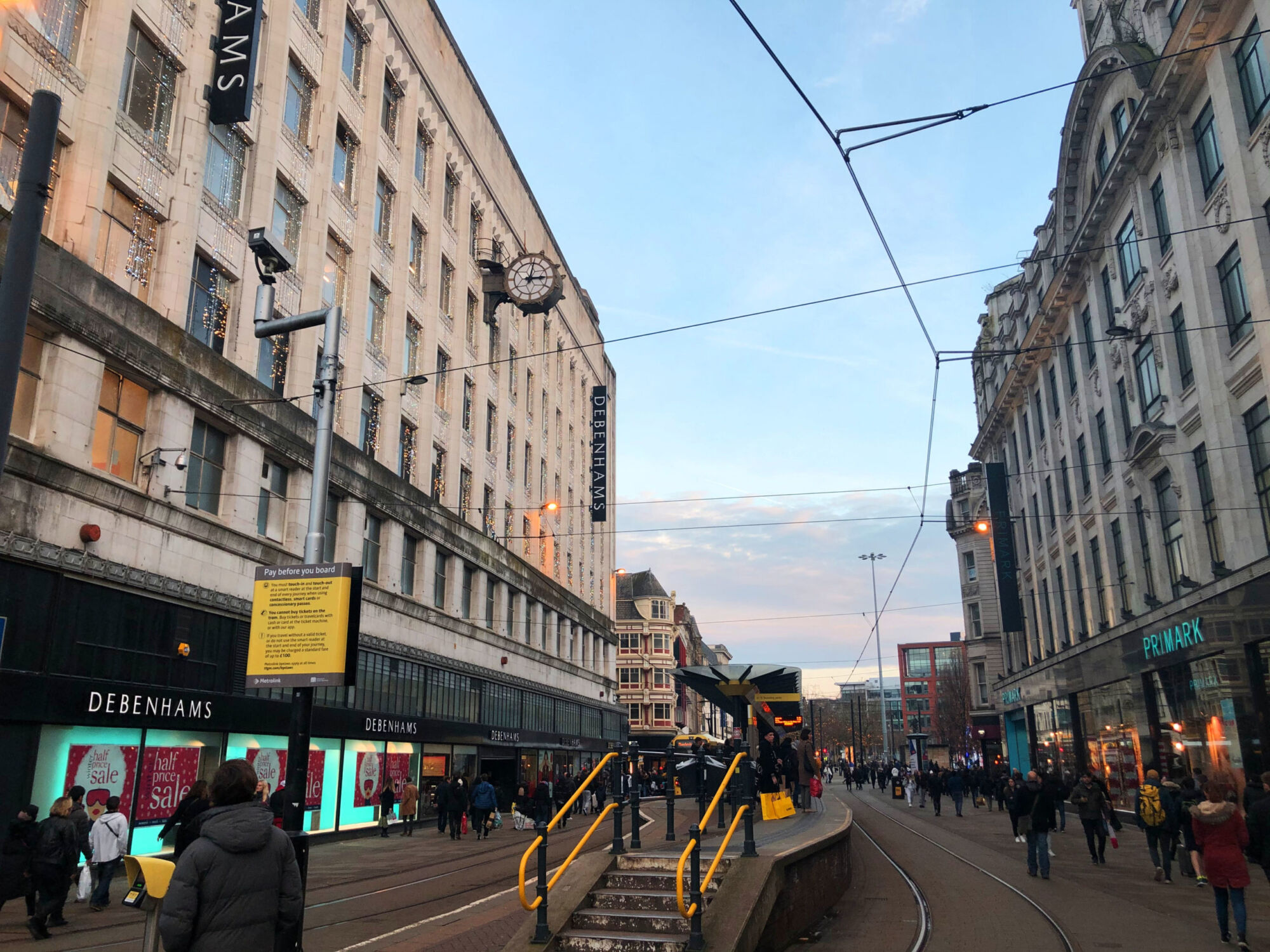 A yellow Metrolink tram at a city-centre stop, shoppers crossing in front of the Debenhams store