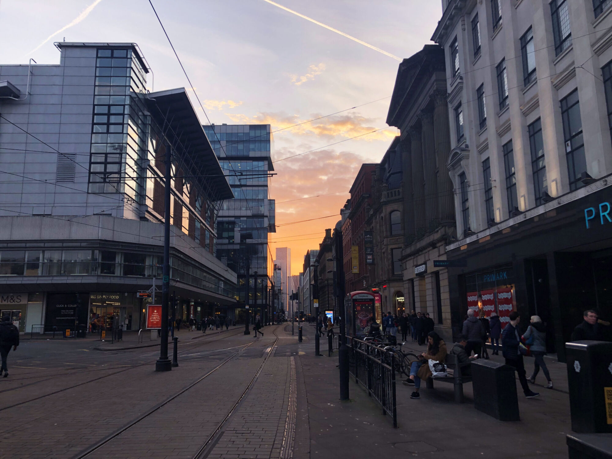 A tram line running down a Manchester street at sunset, the sky pink between the buildings