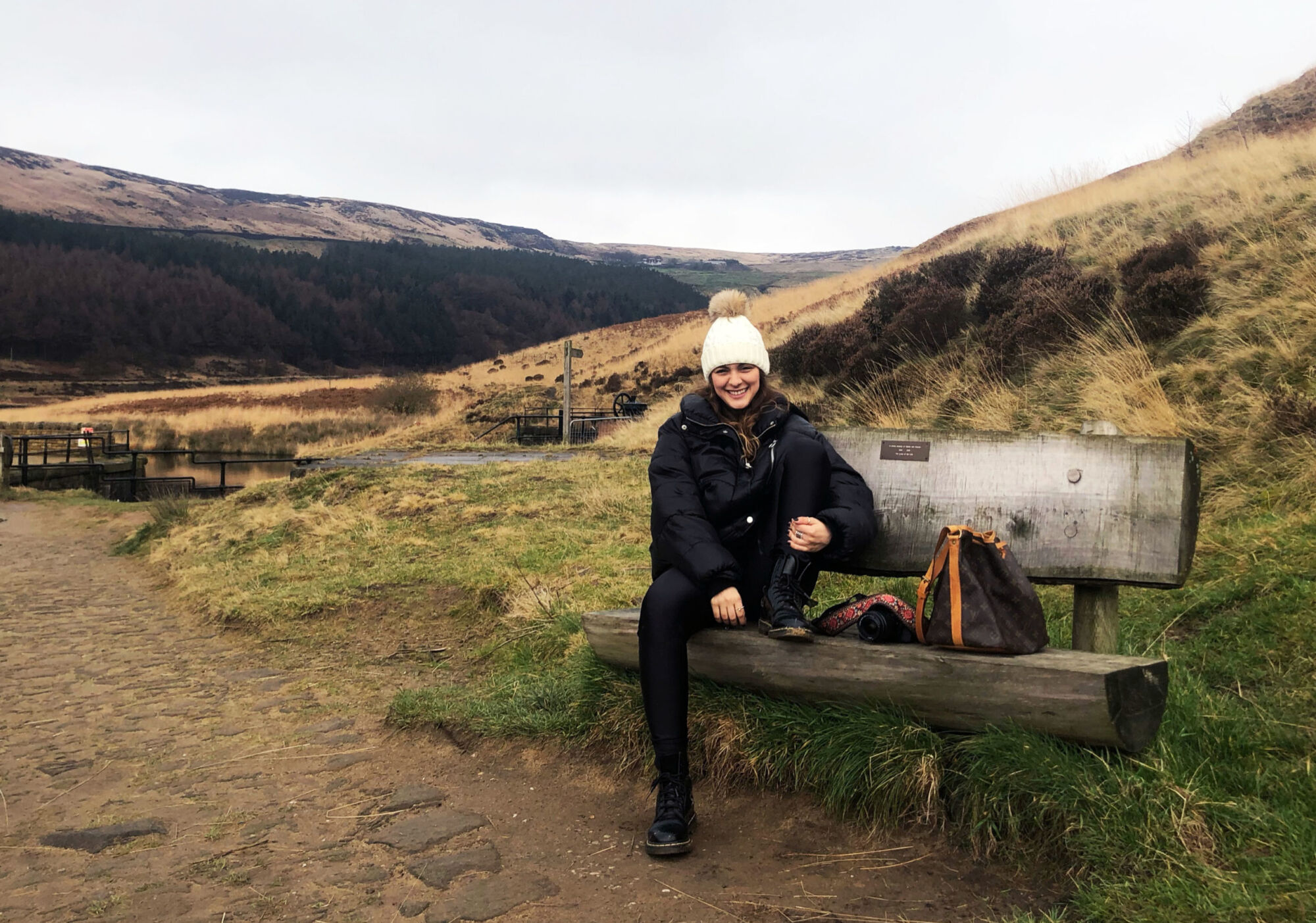 Cata sitting on a wooden bench on the reservoir path in a white bobble hat and long black coat, moorland hills behind her