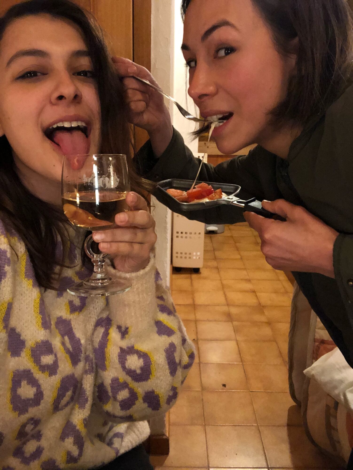 Two women sharing a plate of food indoors, a glass of white wine in hand, both mid-laugh