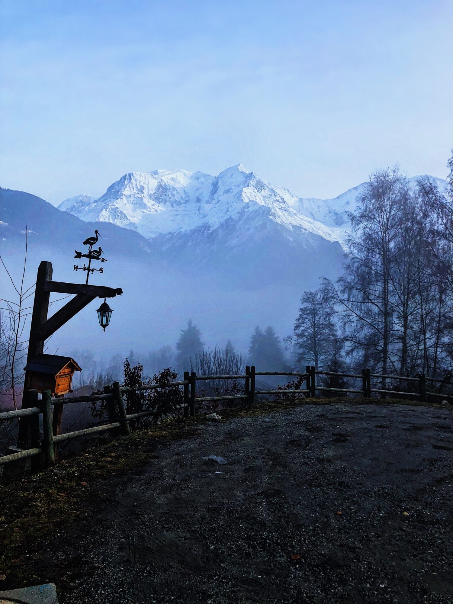 A wrought-iron weathervane on a fence at dusk, mist in the valley and snow-covered peaks catching the last light