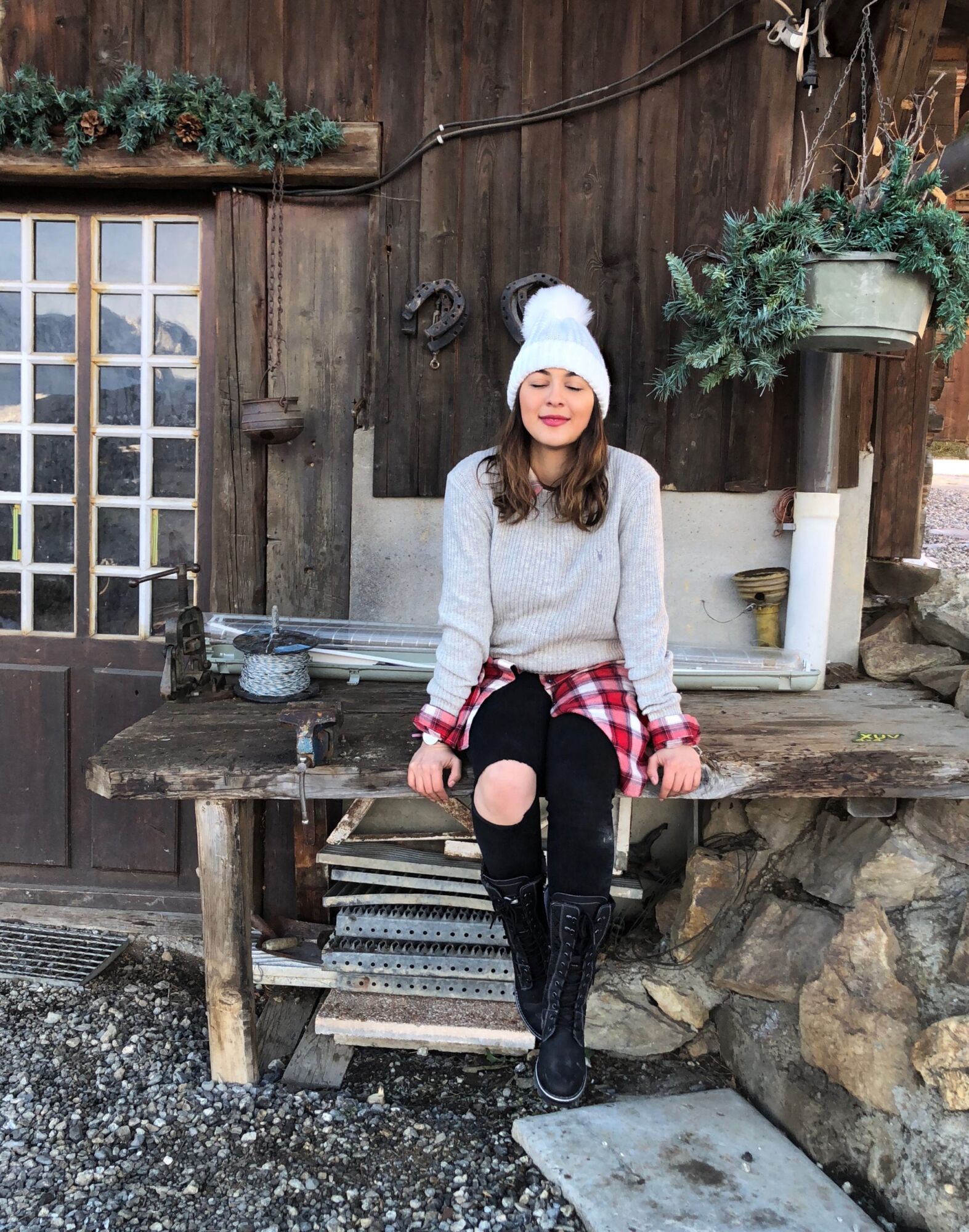 Cata sitting on a wooden bench outside an old chalet in a white bobble hat and black boots, a pine wreath on the wall
