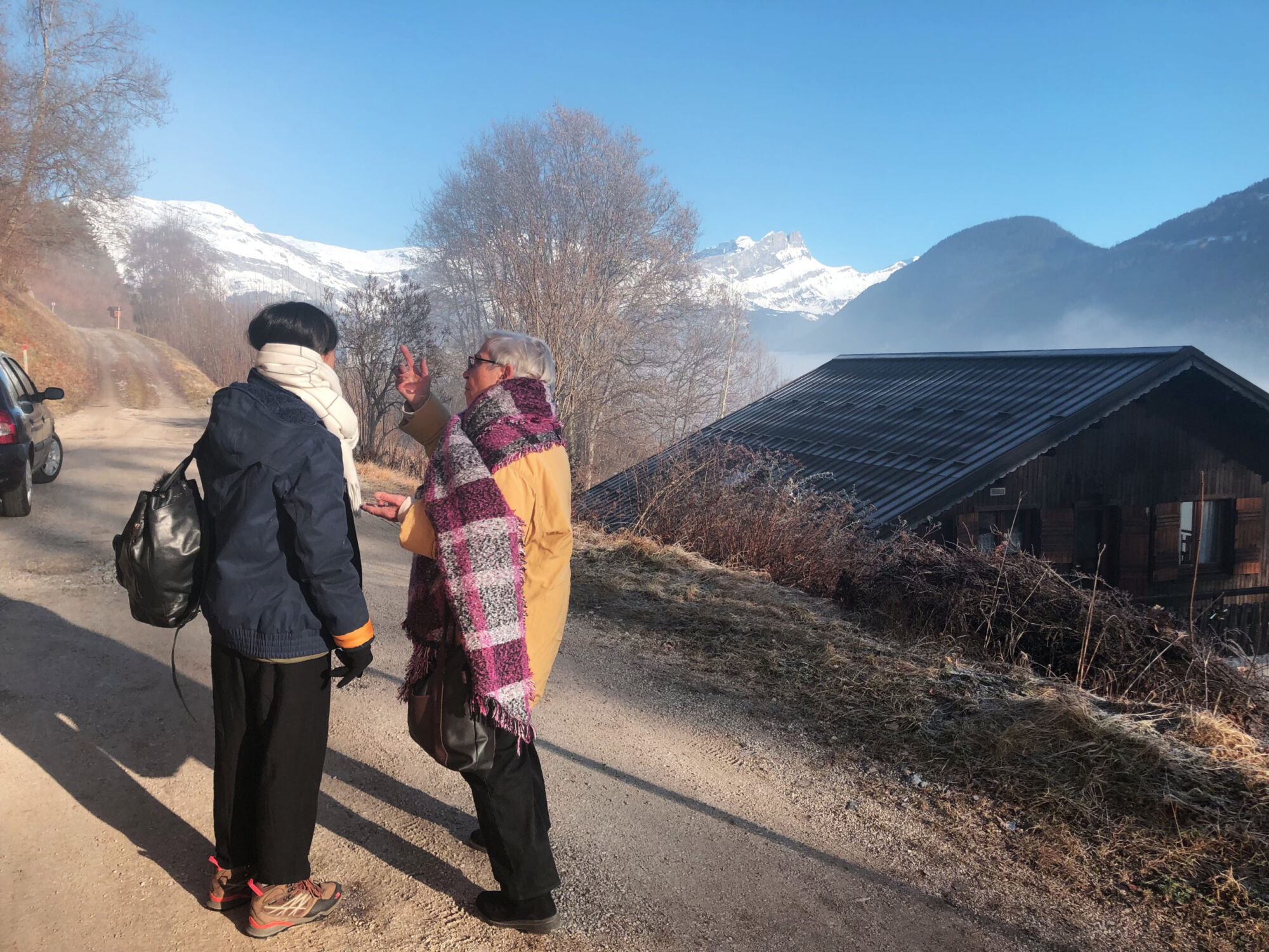 Two people in winter coats and scarves on a mountain road, looking out at snow-covered peaks beside a dark chalet roof