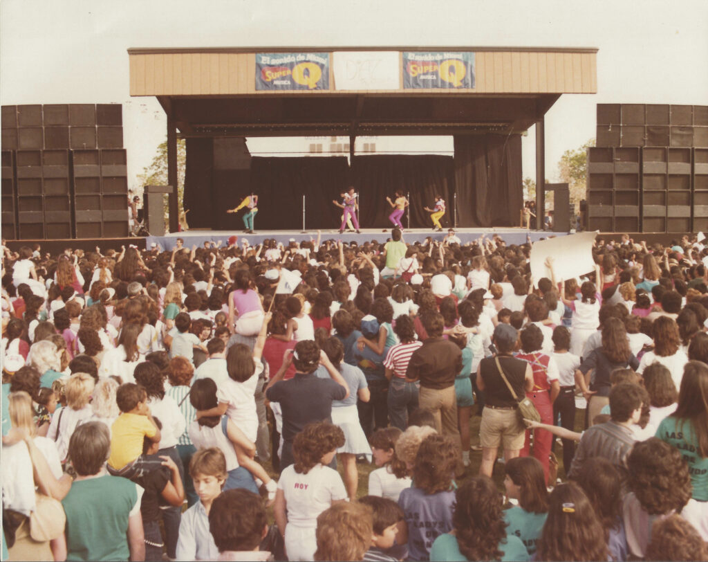 A large crowd watching performers on an outdoor stage, in an old colour photo