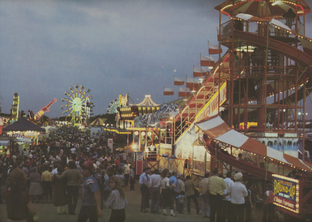 The fairground at dusk with a Ferris wheel and a big slide, crowds below