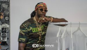 2 Chainz in sunglasses, a bandana and a camouflage T-shirt, leaning on a clothes rail