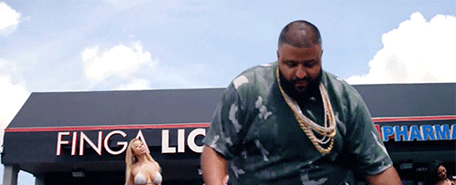 DJ Khaled in a camouflage shirt and gold chains outside a shop with a "Finga Lickin" sign