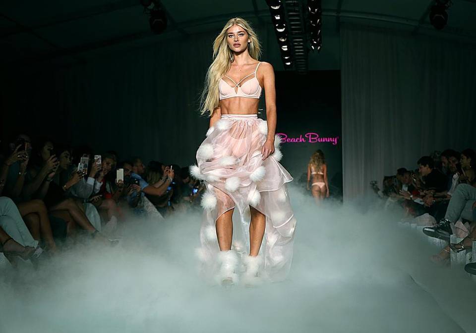 A model in a white bikini and sheer ruffled skirt walking through stage fog at Swim Week