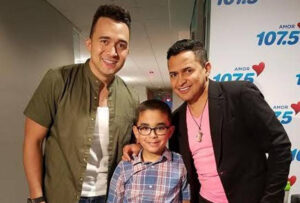 Jorge Celedón and another man posing with a young boy in glasses in front of an Amor 107.5 backdrop