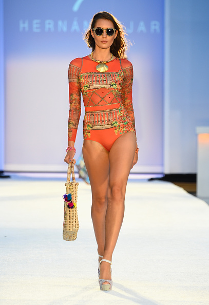 A model in a long-sleeved printed swimsuit carrying a woven bag on the runway
