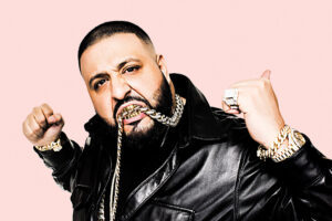 DJ Khaled in a black leather jacket and gold chains, fists raised, against a pink background
