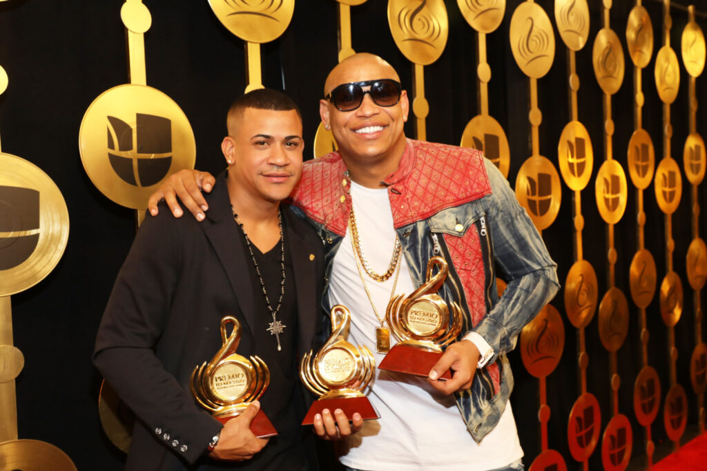 Gente de Zona, two men holding gold Premio Lo Nuestro awards in front of a gold backdrop