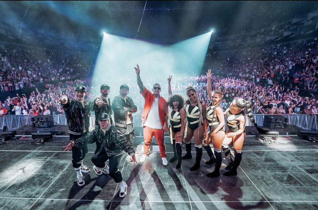 Daddy Yankee and dancers posing on a spotlit stage in front of a packed arena