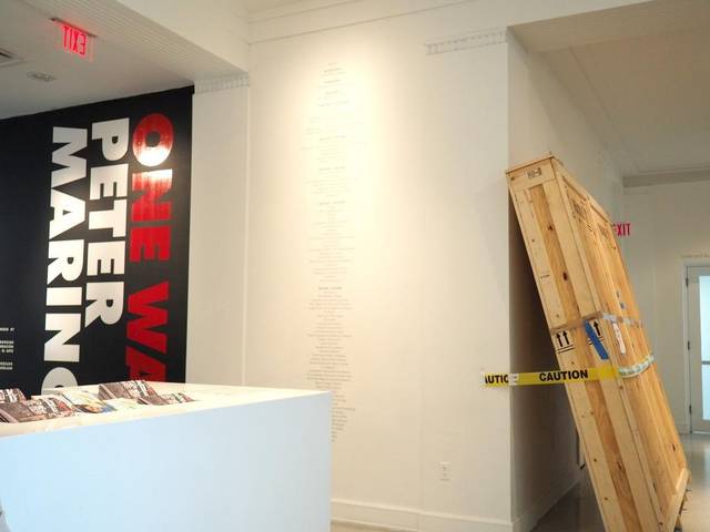 A wooden crate leaning against a wall behind caution tape, beside a Peter Marino exhibition sign