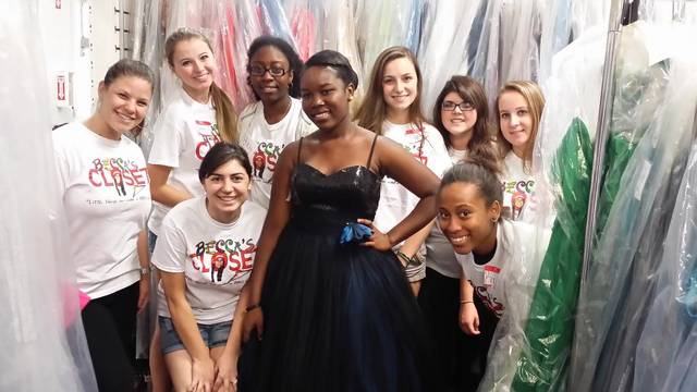 Volunteers in "Closet" T-shirts posing with a girl in a black prom gown among rails of dresses