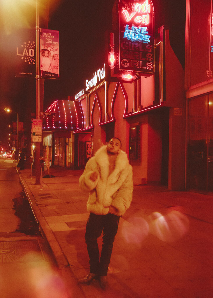 C. Tangana in a cream fur coat on a neon-lit street at night