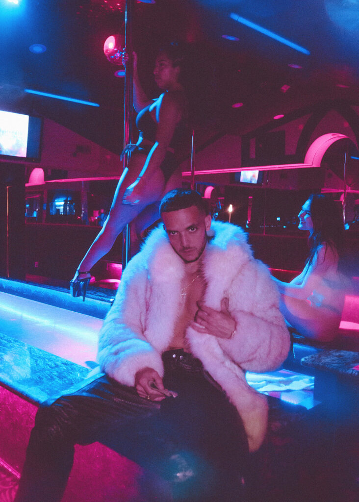 C. Tangana in a white fur coat, sitting in a club lit blue and pink