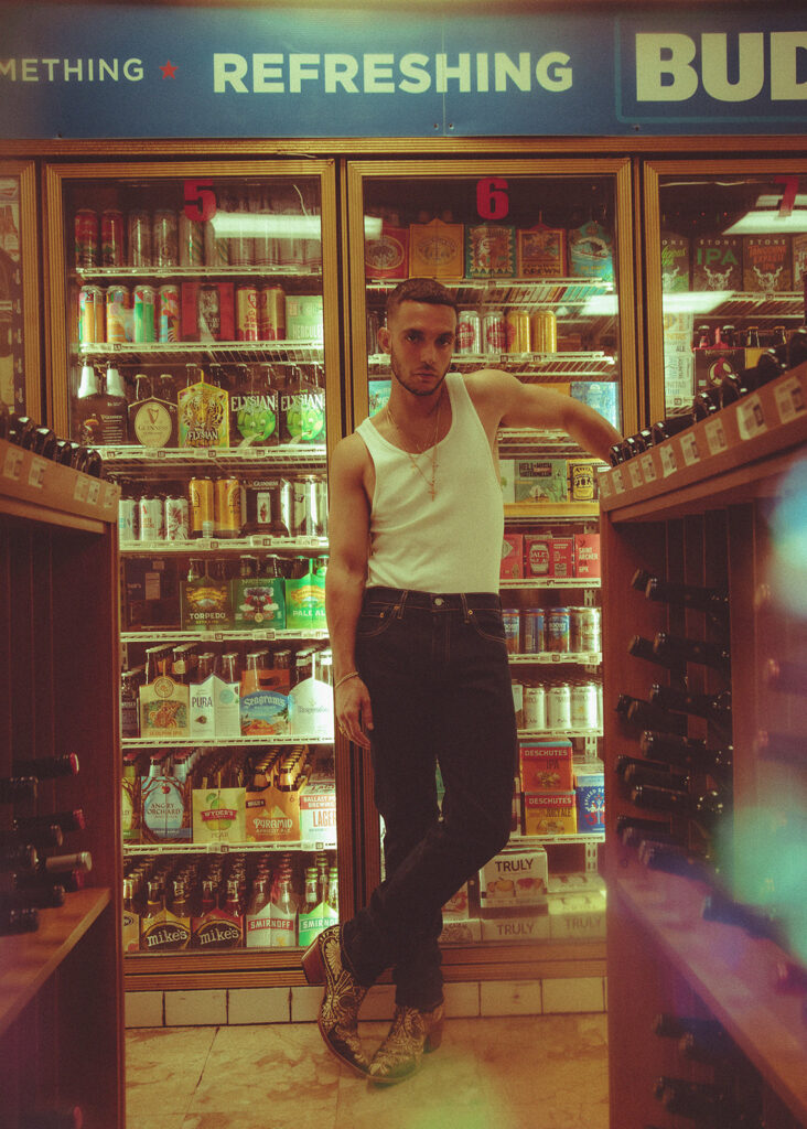 C. Tangana leaning against the drinks fridges inside a liquor store