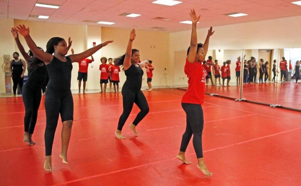 A row of young dancers in red and black stretching their arms up in a mirrored studio