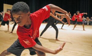 A boy in a red T-shirt in a low dance pose, other dancers behind him in a studio