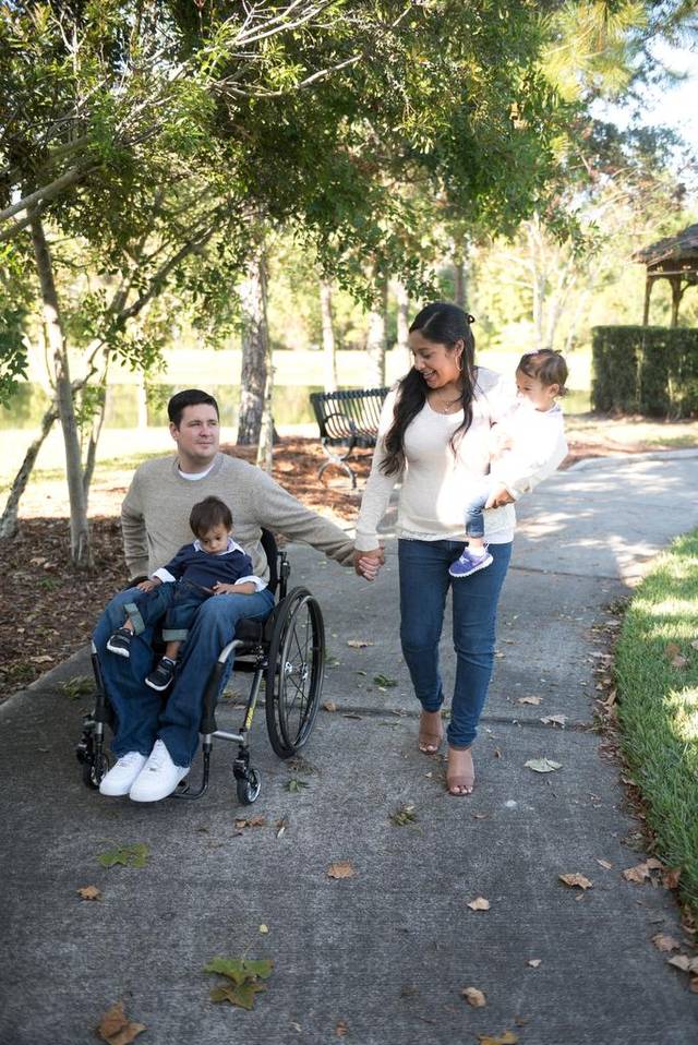 A man in a wheelchair with a child on his lap, a woman walking beside them under trees