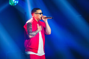 Daddy Yankee in sunglasses and a red jacket, singing into a microphone under blue stage lights