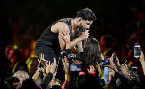 Maluma singing into a microphone at the edge of the stage, fans reaching up with phones