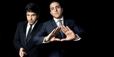 Two men in dark suits, one making a triangle sign with his hands, against a black background