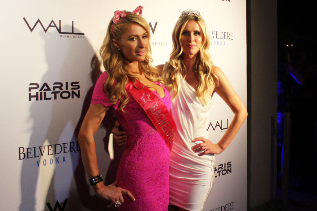 Paris and Nicky Hilton posing together in pink and white in front of a Wall and Paris Hilton backdrop