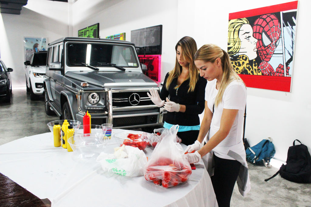 Two women making sandwiches at a table in a gallery, a Mercedes SUV parked behind them
