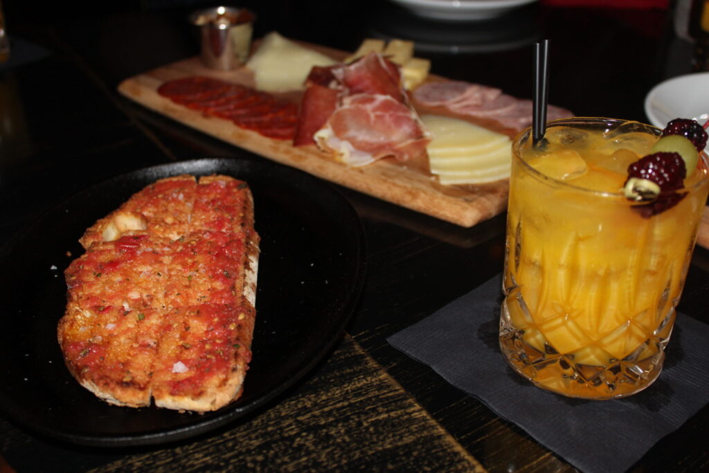 A board of cured meats and cheese beside a flatbread and a cocktail