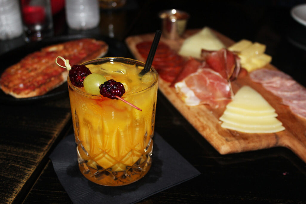 A cocktail beside a board of cured meats and cheese and a flatbread