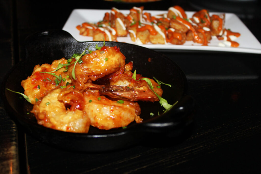 Glazed shrimp on a black plate, with a platter of bruschetta behind
