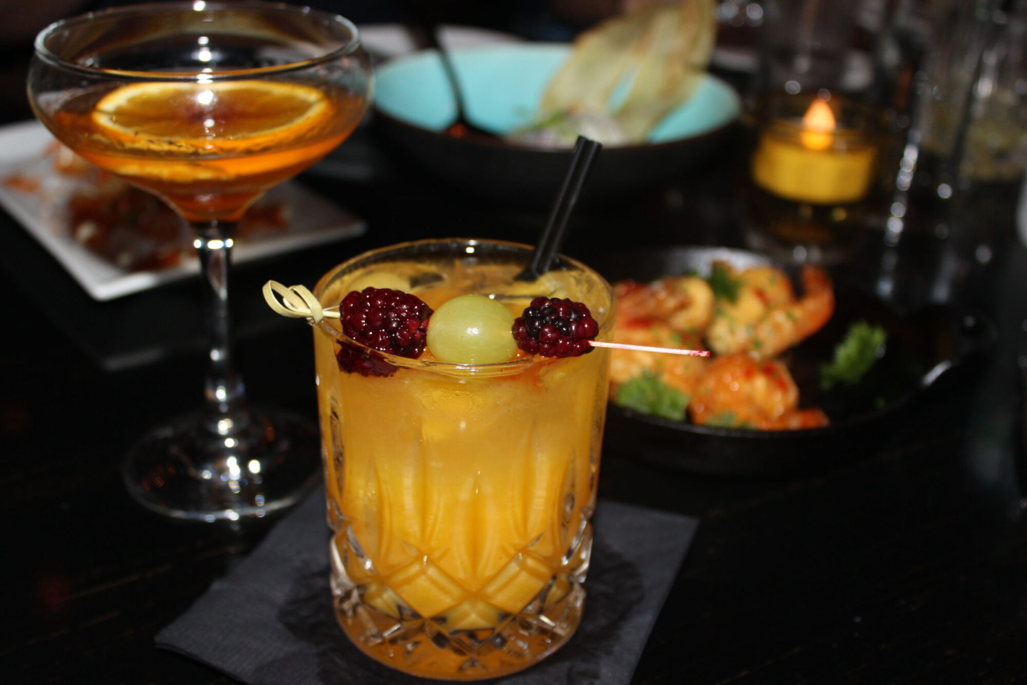 Two cocktails, one garnished with blackberries, on a candlelit bar table