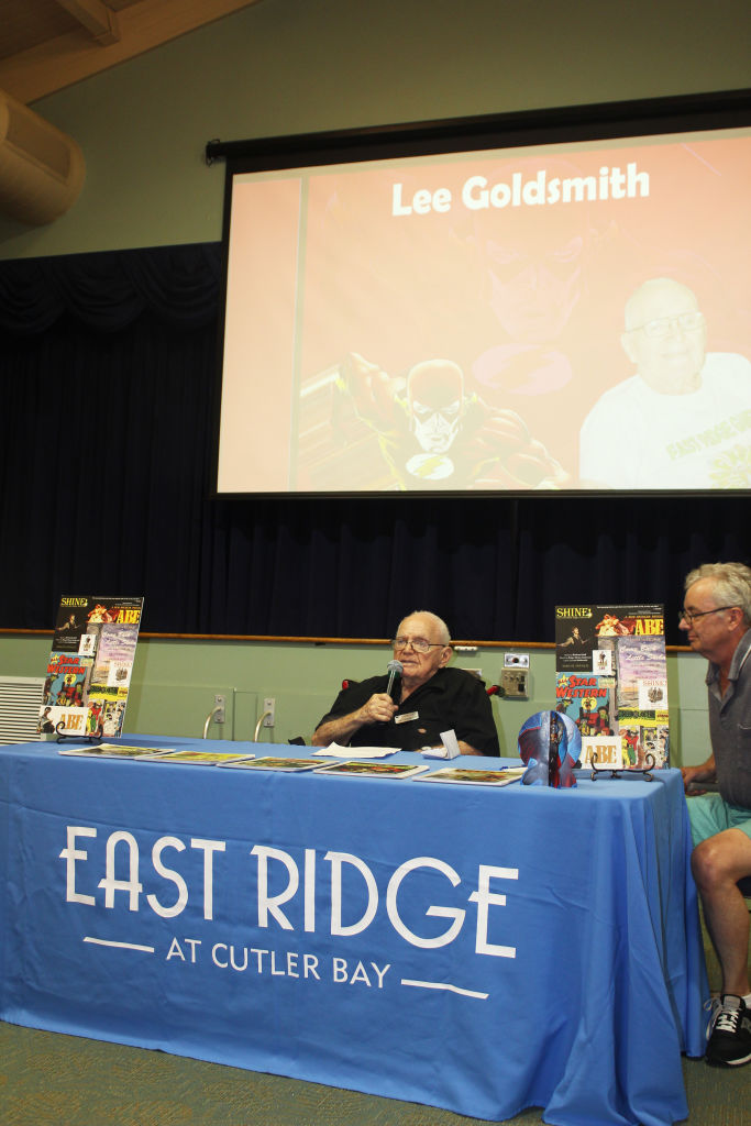 A man at a table with an East Ridge banner, a screen reading "Lee Goldsmith" behind him