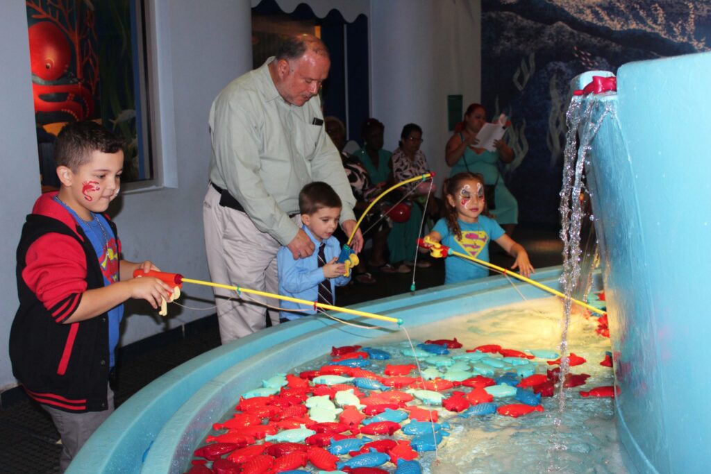 Children fishing with toy rods in a water play area at a children's museum, a man helping them
