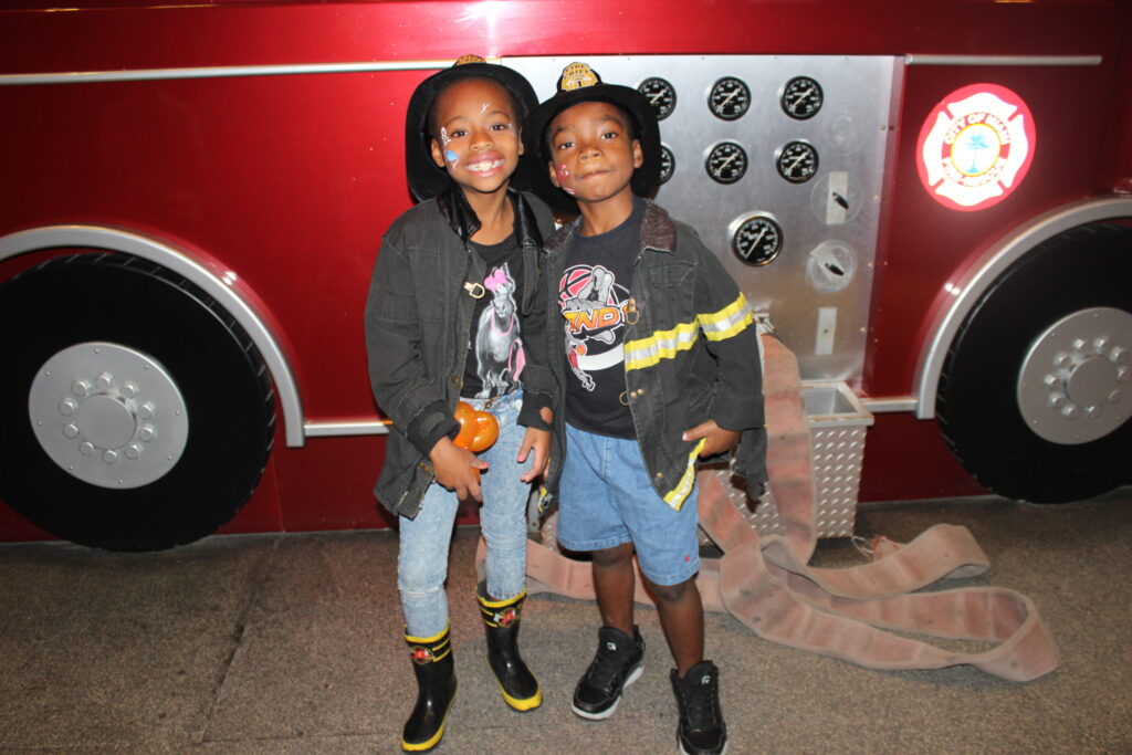 Two children in firefighter outfits posing in front of a red fire truck exhibit