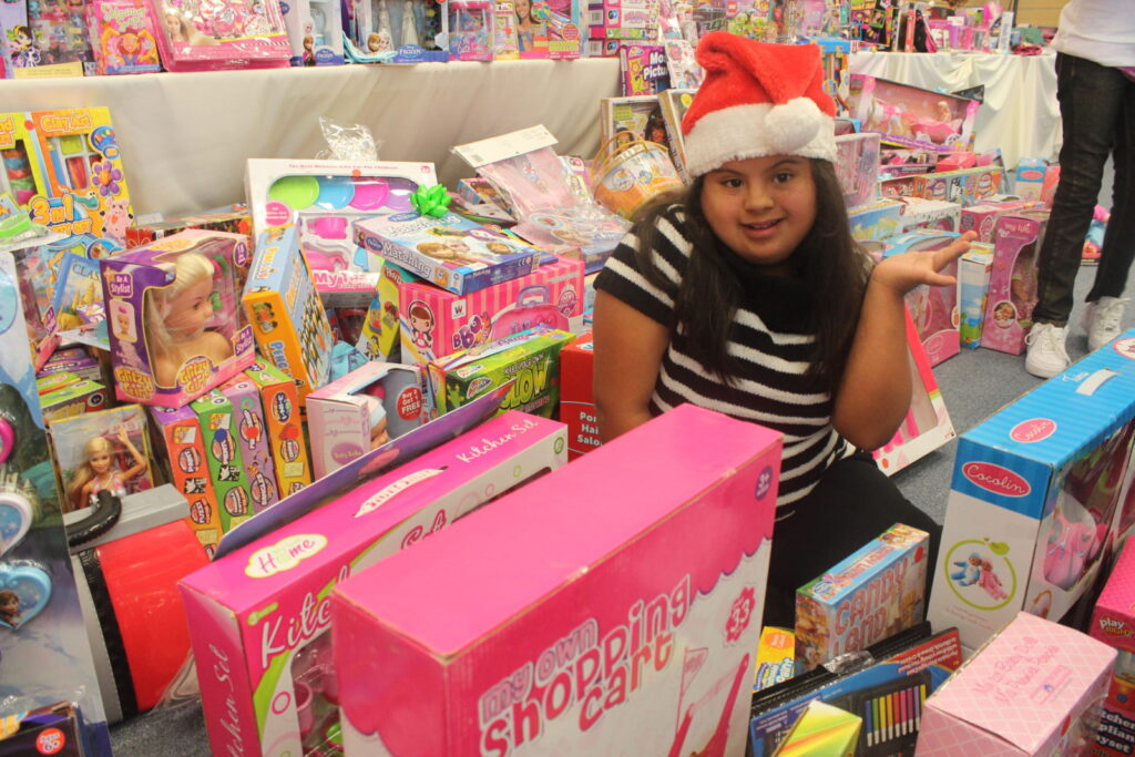 A girl in a Santa hat surrounded by piles of toys in their boxes