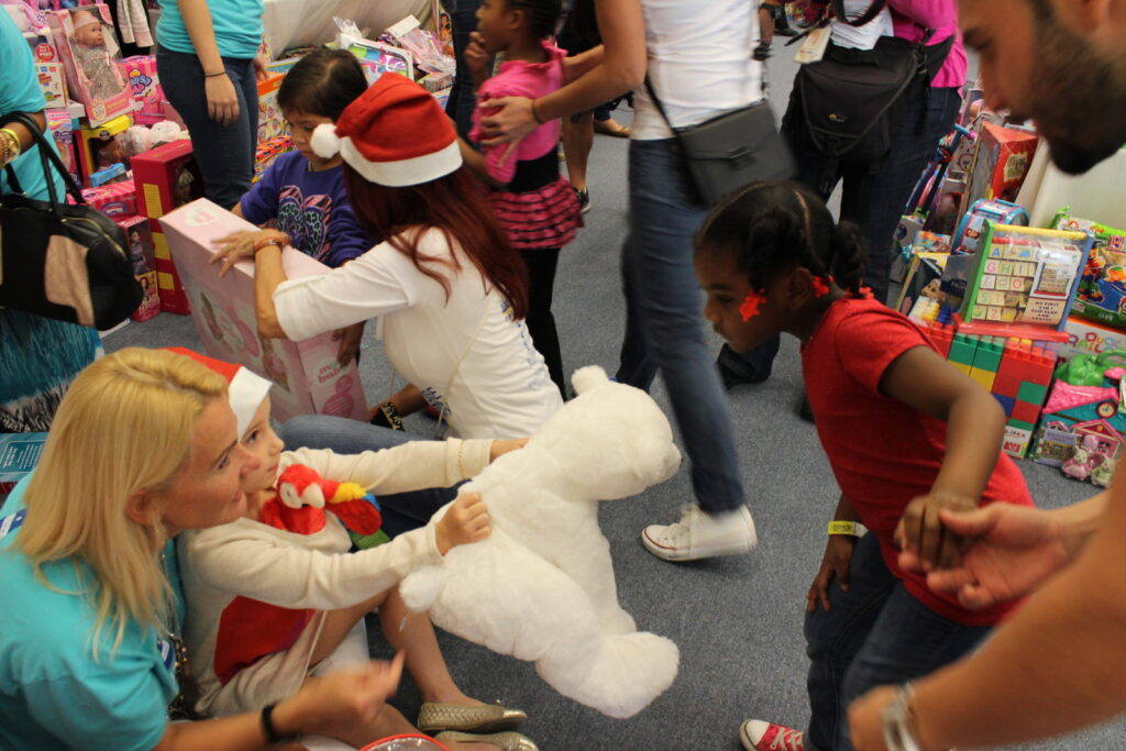 Children in Santa hats and volunteers choosing toys, a girl hugging a white teddy bear
